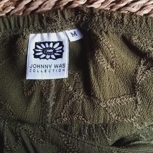 May include: Close-up of olive green fabric with an embroidered leaf pattern. A white tag with the text "JOHNNY WAS COLLECTION" and a medium size tag are visible. The fabric has a textured appearance.
