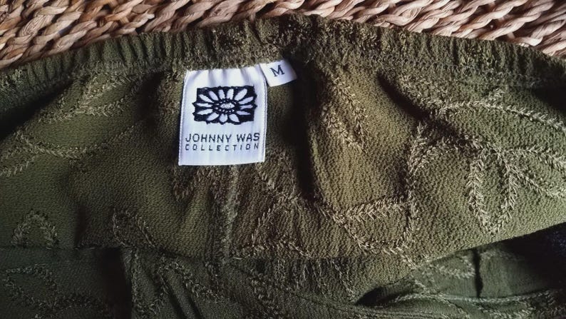 May include: Close-up of olive green fabric with an embroidered leaf pattern. A white tag with the text "JOHNNY WAS COLLECTION" and a medium size tag are visible. The fabric has a textured appearance.