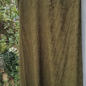 May include: Olive green skirt with embroidered leaf pattern and a velvet hem. The skirt has a button-up front and is hanging on a wooden hanger. The fabric has a textured appearance, and the design is reminiscent of nature.
