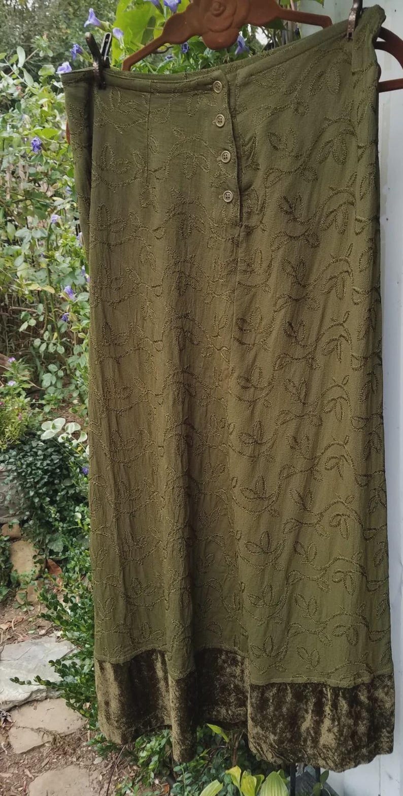 May include: Olive green skirt with embroidered leaf pattern and a velvet hem. The skirt has a button-up front and is hanging on a wooden hanger. The fabric has a textured appearance, and the design is reminiscent of nature.