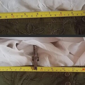 May include: Two sections of a dark green garment with embroidered details are measured with a yellow tape measure. The garment is approximately 13 inches wide. The background is white fabric.