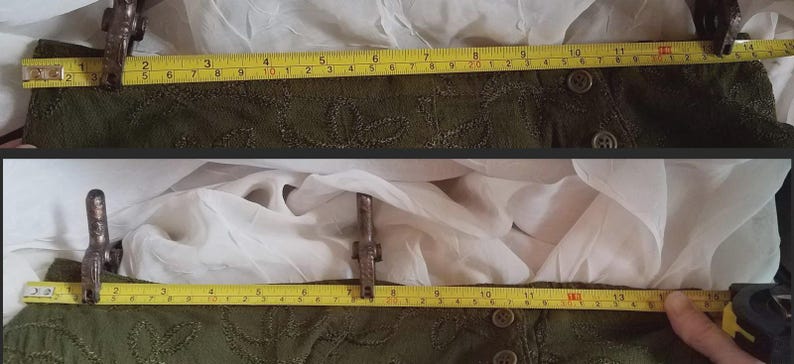 May include: Two sections of a dark green garment with embroidered details are measured with a yellow tape measure. The garment is approximately 13 inches wide. The background is white fabric.