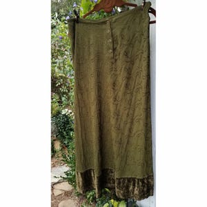 May include: Olive green maxi skirt with a button-up front and embroidered leaf pattern. The skirt has a velvet hem and is hanging on a wooden hanger. The skirt is outdoors, with greenery in the background.