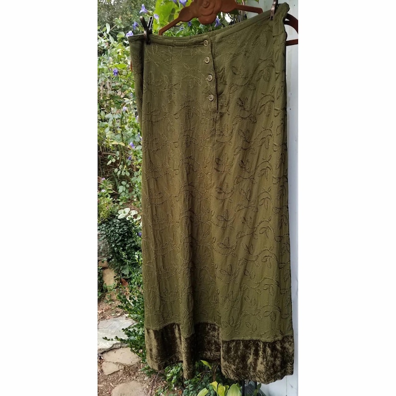 May include: Olive green maxi skirt with a button-up front and embroidered leaf pattern. The skirt has a velvet hem and is hanging on a wooden hanger. The skirt is outdoors, with greenery in the background.