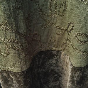 May include: Close-up of olive green fabric with embroidered leaf patterns. The fabric appears to be layered, with a textured top layer and a darker, velvety bottom layer. The stitching is in a matching olive green thread.