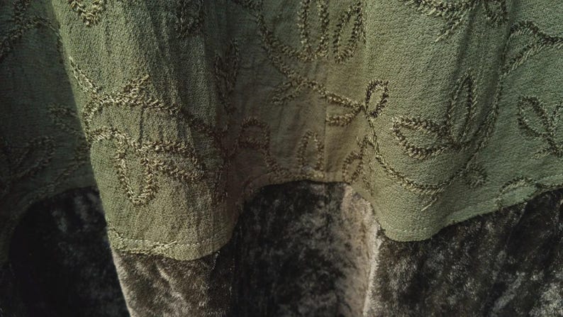 May include: Close-up of olive green fabric with embroidered leaf patterns. The fabric appears to be layered, with a textured top layer and a darker, velvety bottom layer. The stitching is in a matching olive green thread.