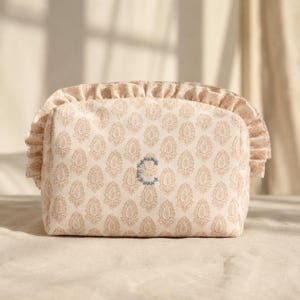 Puede incluir: Un estuche de tela beige con un patrón floral repetitivo de color marrón claro y un borde con volantes. El centro del estuche está bordado con una letra C azul en estilo de punto de cruz, aportando un acento decorativo al diseño.