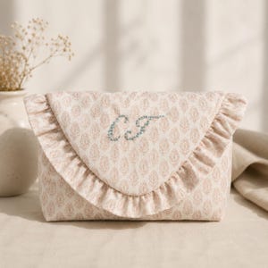 Puede incluir: Un estuche de tela con un estampado floral en beige claro y naranja suave. La solapa estilo sobre está adornada con un borde de volantes y bordada con las iniciales azules CJ en una fuente cursiva en la parte central delantera del objeto.
