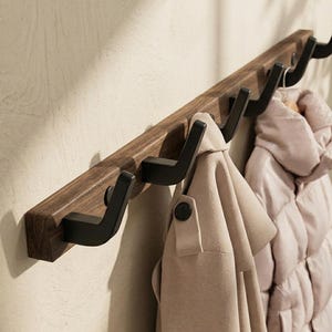 Könnte beinhalten: Eine Wandgarderobe mit einem horizontalen, dunkelbraunen Holzbrett und fünf mattschwarzen Metallhaken. Ein beigefarbener Trenchcoat und eine hellrosa Steppjacke hängen an den Haken und zeigen das funktionale Design.