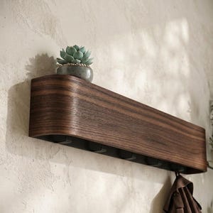 Könnte beinhalten: Dunkles Holz-Wandregal mit geschwungenem Design, mit einer kleinen Topf-Sukkulente. Das Regal hat mehrere Haken darunter zum Aufhängen von Gegenständen. Eine braune Jacke hängt an einem der Haken. Das Regal ist an einer strukturierten Wand befestigt.