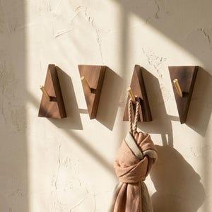 Pode incluir: Quatro ganchos de parede de madeira com pinos de latão em um design triangular. Um gancho segura um cachecol amarrado. Os ganchos são feitos de madeira escura e são montados em uma parede texturizada esbranquiçada. O cachecol é uma mistura de rosa e bege.
