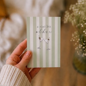 May include: A rectangular greeting card featuring a vertical sage green and white striped pattern. The text "a tini bit older" is printed in black, accompanied by a line-art illustration of two martini glasses with olives on toothpicks.