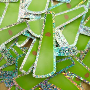May include: A collection of die-cut stickers featuring a bright green, elongated shape with a holographic border. Each sticker displays a small brown oval accent and the lowercase black text "they left." printed on the surface.