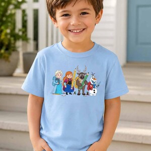 Puede incluir: Una camiseta de manga corta de color azul claro con un estampado gr&aacute;fico de cuatro personajes de dibujos animados de Frozen, incluidos Elsa, Anna, Sven y Olaf, todos representados con material escolar como mochilas, l&aacute;pices y libros.