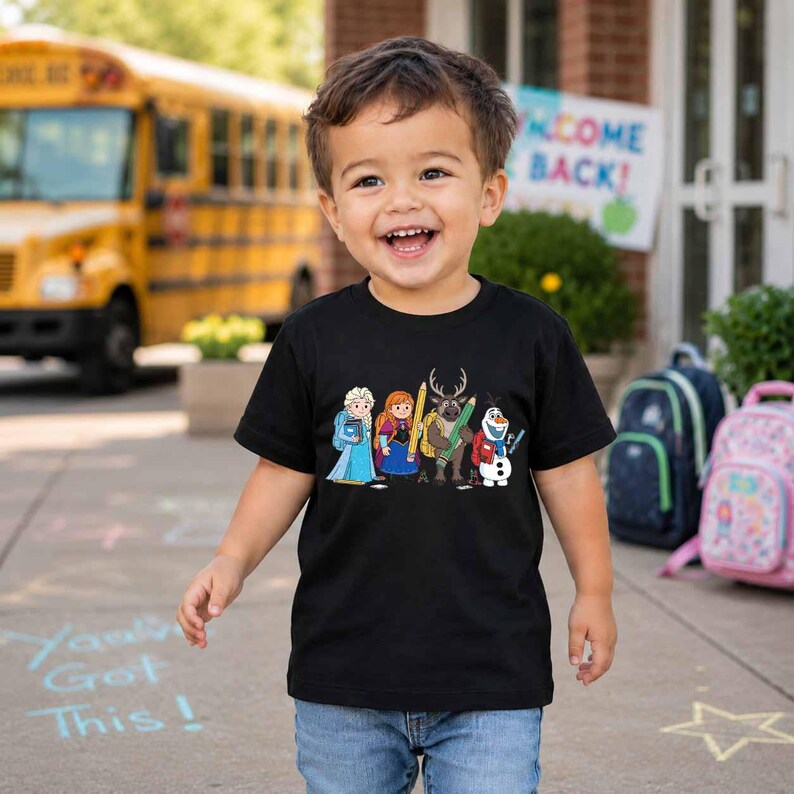 Puede incluir: Una camiseta negra de manga corta con un estampado gr&aacute;fico de los personajes de dibujos animados Elsa, Anna, Sven y Olaf de Frozen, cada uno con material escolar como l&aacute;pices y libros, sobre un fondo de tela negra lisa.