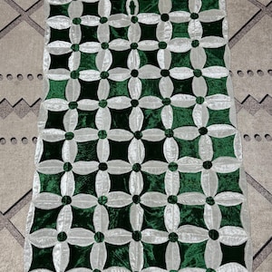 May include: A rectangular, decorative table runner with a repeating floral pattern. The design features dark green and silver-white velvet-like fabric. The runner's pattern is a series of four-petal flowers and square shapes.