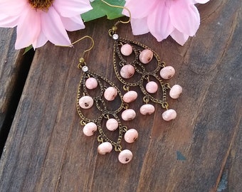 May include: A pair of dangle earrings with a delicate, ornate design. The earrings feature a teardrop-shaped frame with a cascading row of pink beads. The beads are made of a light pink stone with a subtle marbled pattern.