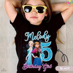 Może przedstawiać: Czarny t-shirt z napisem "Melody" w niebieskim skrypcie, cyfrą "5" w błyszczącym niebieskim i słowami "Birthday Girl" w różowym skrypcie. Koszulka zawiera również wizerunki dwóch postaci z kreskówek.