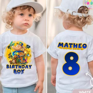 May include: A white t-shirt with a colorful cartoon graphic and the text "BIRTHDAY BOY" in blue and yellow. The back of the shirt has the name "MATHEO" and the number "8" in blue and yellow. The child is wearing a white baseball cap.