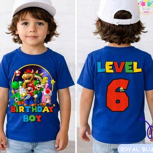 May include: Royal blue t-shirt featuring a colorful graphic of video game characters and the text "BIRTHDAY BOY" on the front. The back of the shirt displays "LEVEL 6" in bold, colorful letters. The child is wearing a white baseball cap.