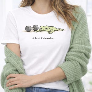 May include: White t-shirt featuring a cartoon frog lying next to a dumbbell. The frog has a bored expression. The text "at least i showed up" is printed below the frog and dumbbell.