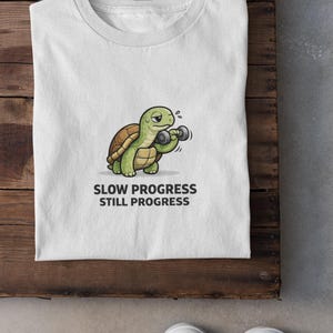 Op de afbeelding: Wit T-shirt met een cartoon schildpad die gewichten heft. De schildpad is groen en bruin, met zweetdruppels. De tekst "SLOW PROGRESS STILL PROGRESS" staat eronder. Het shirt ligt op een houten oppervlak.