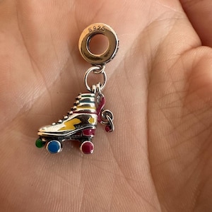 May include: A silver roller skate charm with colourful accents. The skate features yellow, red, green, and blue details. The charm is attached to a silver ring with the marking "S925". A pink ribbon is attached to the skate.