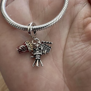 May include: Silver charm with movie-themed pendants. The charm features a popcorn bucket, a film camera, and a film reel. The word "MOVIE" is inscribed on the charm. The charm is attached to a silver bracelet.