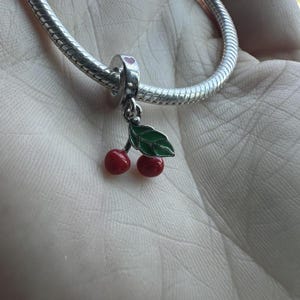 Puede incluir: Un charm de plata con un diseño de cereza. El charm presenta dos cerezas rojas con una hoja verde y un lazo plateado con un corazón rosa. El charm está unido a una pulsera de plata.