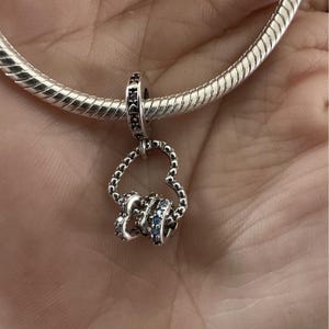 May include: A silver charm with a heart-shaped design and three dangling rings. The charm features small blue and clear gemstones. The charm is attached to a silver bracelet.