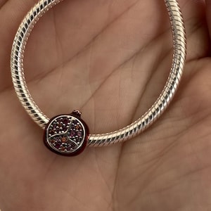 Könnte beinhalten: Ein silbernes Armband mit einem Anhänger mit einem roten Granatapfel-Design. Der Anhänger ist mit kleinen, farbigen Edelsteinen verziert. Das Armband hat einen strukturierten Schlangenketten-Stil.