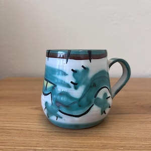 Op de afbeelding: Een keramische mok met een witte basis voorzien van abstracte blauwgroene en groene geschilderde wervelingen, geaccentueerd door dunne handgeschilderde zwarte lijnen en een donkerbruine rand. De mok heeft een ronde vorm en een gebogen handvat.