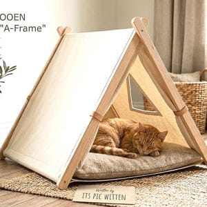 May include: A minimalist A-frame cat house made of light wood and canvas. An orange tabby cat is sleeping inside on a beige cushion. The text "DIY WOEN Minimalist A-Frame" is visible.