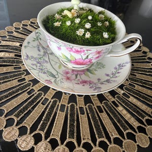 May include: A floral teacup and saucer filled with green moss, miniature white flowers, and a small figurine. The teacup rests on a gold and black decorative placemat. A charming, garden-inspired arrangement.