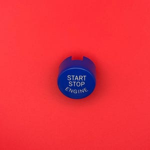 May include: A blue engine start-stop button with white text on a red background. The button is circular with a small notch at the top. The text reads "START STOP ENGINE".