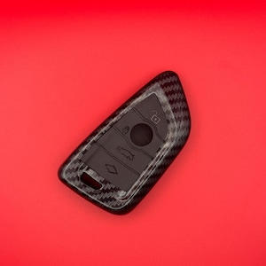 Pode incluir: Um chaveiro de carro preto com uma capa texturizada em fibra de carbono. O chaveiro tem vários botões e um design de fechadura. O fundo é vermelho sólido.