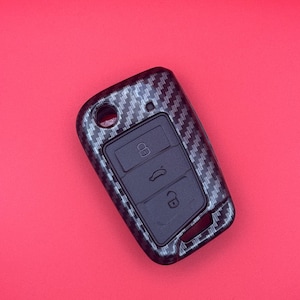 Op de afbeelding: Een autosleutelhanger met een koolstofvezelpatroon. De sleutelhanger heeft drie zwarte knoppen met pictogrammen voor vergrendelen, ontgrendelen en het openen van de kofferbak. De achtergrond is effen rood.
