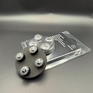Op de afbeelding: Een set zwarte en zilveren ventieldopjes met het Mercedes-Benz logo. De doppen worden gepresenteerd op een zwart schuimrubberen pad en in een transparante plastic verpakking. De doppen hebben een cirkelvormig ontwerp met een zilveren embleem.