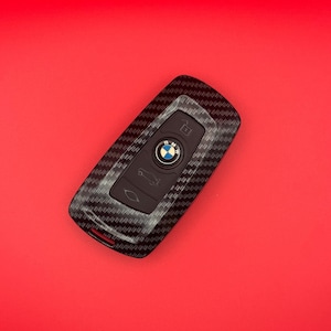 May include: A black and grey carbon fibre car key fob cover with the BMW logo on a red background. The key fob has several buttons and a key ring hole.