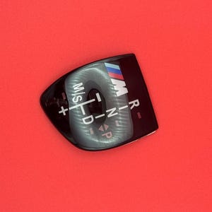 Peut inclure: Cache de levier de vitesses noir avec texte blanc et logo bleu, rouge et blanc. Le cache porte les lettres "M/S", "D", "R", "N" et "P", ainsi que des symboles plus et moins.