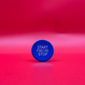 Op de afbeelding: Een blauwe ronde knop met de woorden "START ENGINE STOP" in witte tekst. De knop is gecentreerd op een felrode achtergrond.