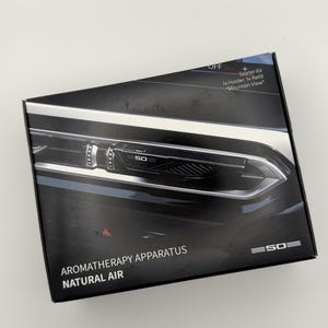 Puede incluir: Una caja de producto rectangular negra para un aparato de aromaterapia. La caja presenta un gráfico de una rejilla de ventilación de coche con el número 50, el texto "AROMATHERAPY APPARATUS NATURAL AIR" y "Starter Kit 1x Holder 1x Refill Mountain View".