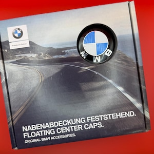 以下が含まれることがあります： 沿岸道路の画像、BMWロゴ、および「NABENABDECKUNG FESTSTEHEND. FLOATING CENTER CAPS. ORIGINAL BMW ACCESSORIES.」というテキストが印刷された正方形のBMW製品ボックス。青と白のBMWロゴが入った円形のフローティングセンターキャップが見えます。