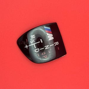 May include: A black gear shift cover with white and silver lettering and the BMW M logo in blue, red, and white. The cover has the letters "M/S", "D", "R", "N", and "P" on it, along with plus and minus symbols.