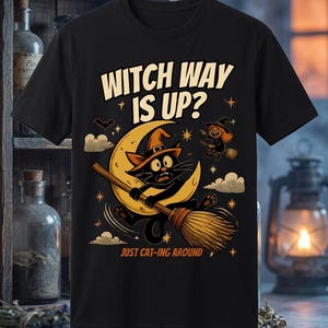 Può includere: Una t-shirt nera con la grafica di un gatto nero con cappello da strega a cavallo di una scopa davanti a una luna crescente gialla. Il testo recita "WITCH WAY IS UP?" e "JUST CAT-ING AROUND" con piccole stelle, nuvole e una strega.