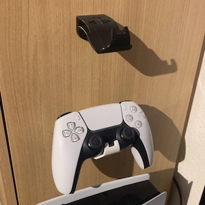 Puede incluir: Un mando de PlayStation 5 blanco y negro con un soporte negro. El mando tiene un diseño simétrico con un panel táctil y botones. El soporte es negro y está diseñado para sujetar el mando.
