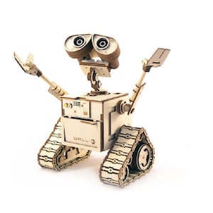 May include: A wooden model of the WALL-E robot, featuring a box-shaped body, articulated arms, and tank treads. The robot has large, expressive eyes and the name "WALL-E" is printed on the front. The model is made of light-colored wood.