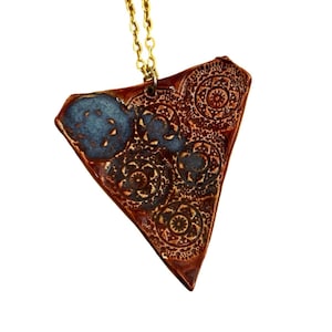 May include: A copper-colored pendant necklace with a gold chain. The pendant is a triangular shape with a blue and brown design. The pendant has a textured surface with circular patterns.