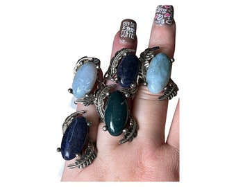 May include: A hand displays six gemstone rings. Each ring features an oval-shaped stone in shades of blue, green, and dark blue, set in a silver-colored metal with a leaf design. The nails have coffee-themed nail art.