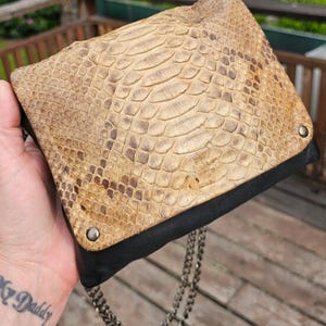 Op de afbeelding: Een beige en bruine clutch met slangenpatroon, een zwarte basis en een zilveren kettingriem. De clutch is rechthoekig met een klepsluiting en twee zilveren klinknagels. Het slangenpatroon heeft een getextureerde uitstraling.